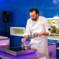 Chef in a white coat stirs a sauce in a stainless steel pot on an induction cooktop during a cooking demonstration, blue backdrop behind him.